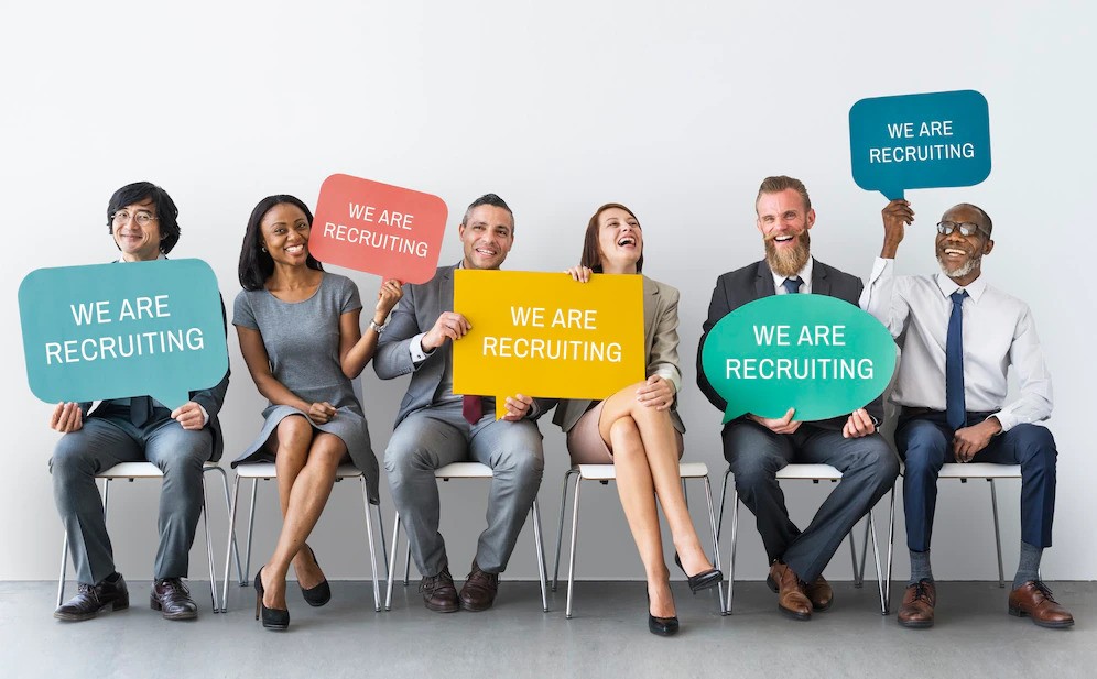 People sitting on chairs holding signs that read We Are Recruiting. This image is to depict real estate brokers looking to hire Real Estate Agents