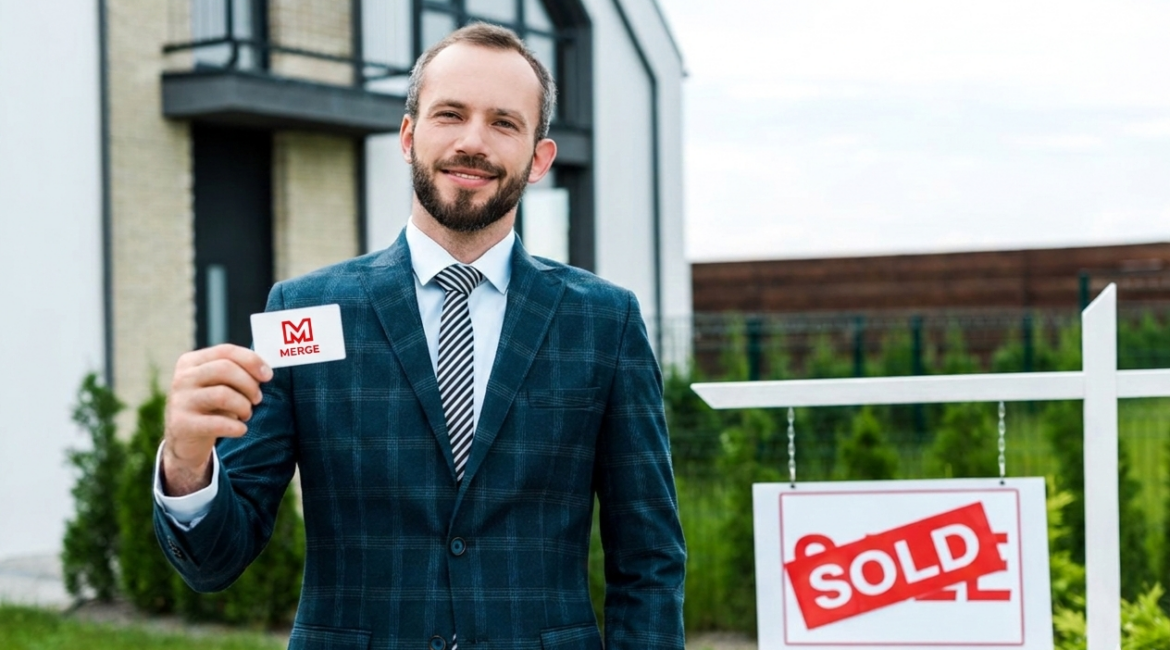 Merge Realty real estate agent holding a merge realy business card infront of a house the he sold.